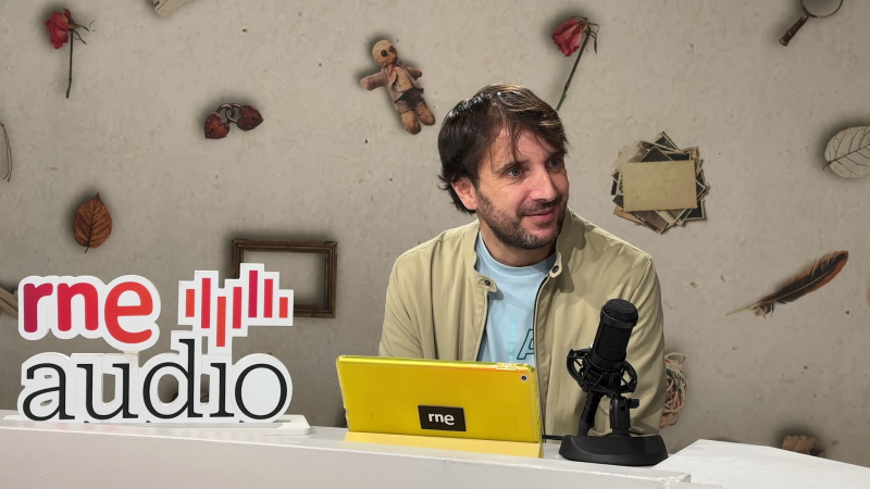 Un hombre con barba y camisa azul clara está sentado frente a una mesa con un micrófono y una tablet. En la pared detrás de él, se observan objetos decorativos como una rosa seca y una muñeca de trapo, junto al logo de "rne audio".