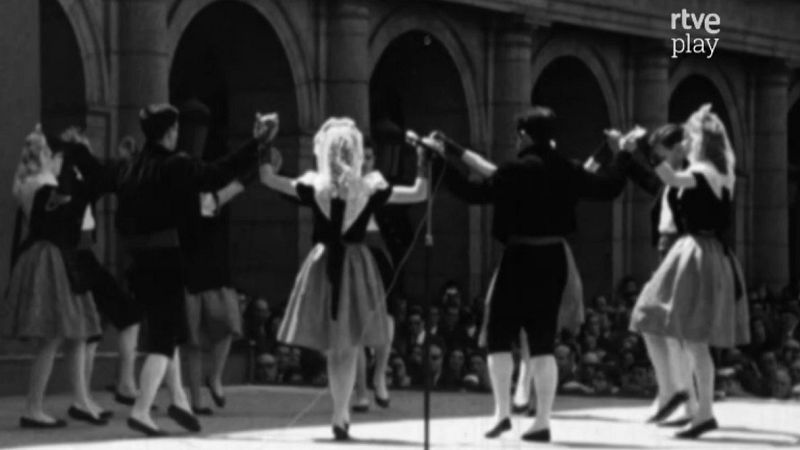 Danza tradicional en blanco y negro con bailarines en círculo, arquitectura clásica y público. Se observa un micrófono y el logo "rtve play".
