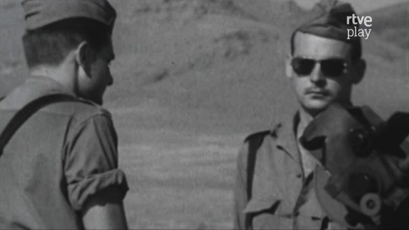 Soldados en un entorno árido, uno de frente con gafas de sol y el otro de espaldas. Fotografía histórica en blanco y negro con logo de RTVE Play.