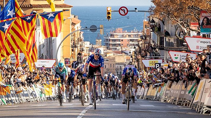 Un pelotón de ciclistas compite en una calle urbana, con espectadores a ambos lados y el mar Mediterráneo como telón de fondo. Se observan banderas y carteles publicitarios.