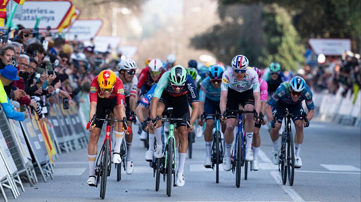 En una carrera de ciclismo, un grupo de competidores, con uniformes de diferentes colores y logotipos, pedalean por una carretera. Se distingue un ciclista con maillot rojo y amarillo, otro con uno negro y verde con la palabra \"Adecco\", y otros con maillots azules y blancos, mientras el público observa la competición.