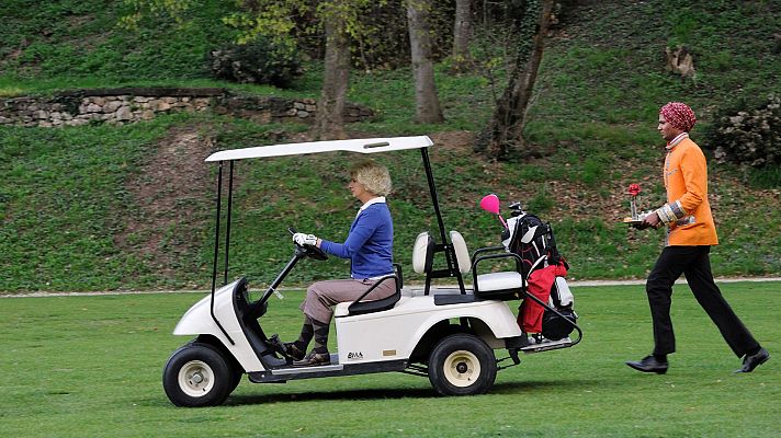 Una mujer rubia conduce un carro de golf blanco, mientras un hombre con turbante y traje colorido camina con una bandeja y una rosa roja en un campo verde con árboles.