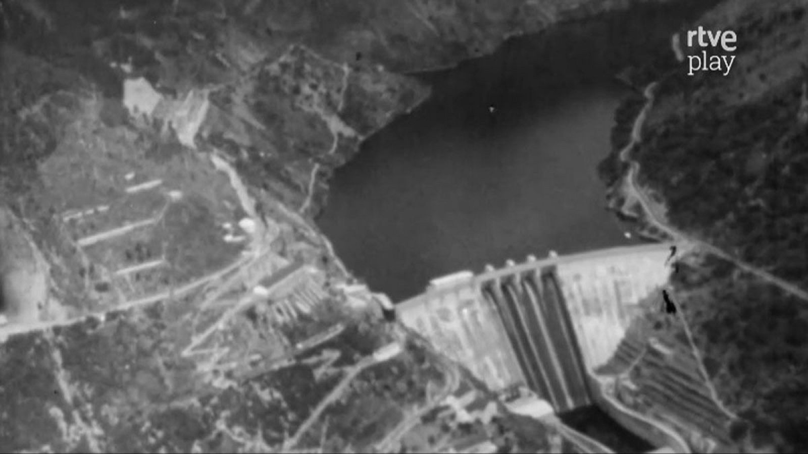 Una fotografía aérea en blanco y negro muestra una presa y un embalse. Se observa la estructura de la presa y el agua fluyendo, con el logo de RTVE Play en la esquina superior derecha.