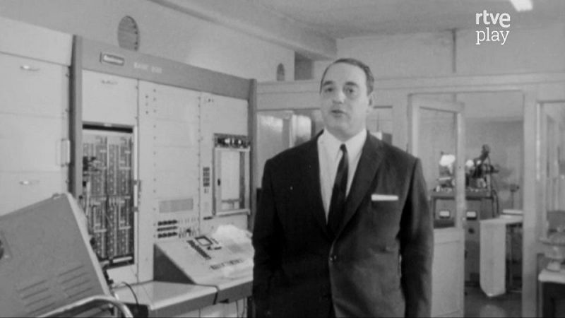 Un hombre con traje y corbata se encuentra frente a un ordenador antiguo en un laboratorio, con paneles y luces, evocando una est�tica de mediados del siglo XX. La imagen pertenece a una serie documental.