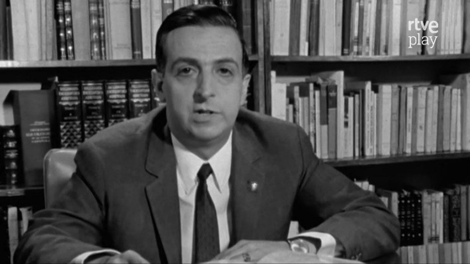 Un hombre de mediana edad, con traje y corbata, está sentado frente a un escritorio, posiblemente durante una entrevista. Se aprecia un reloj en su muñeca y un anillo en su mano, mientras que estanterías llenas de libros se vislumbran detrás de él.