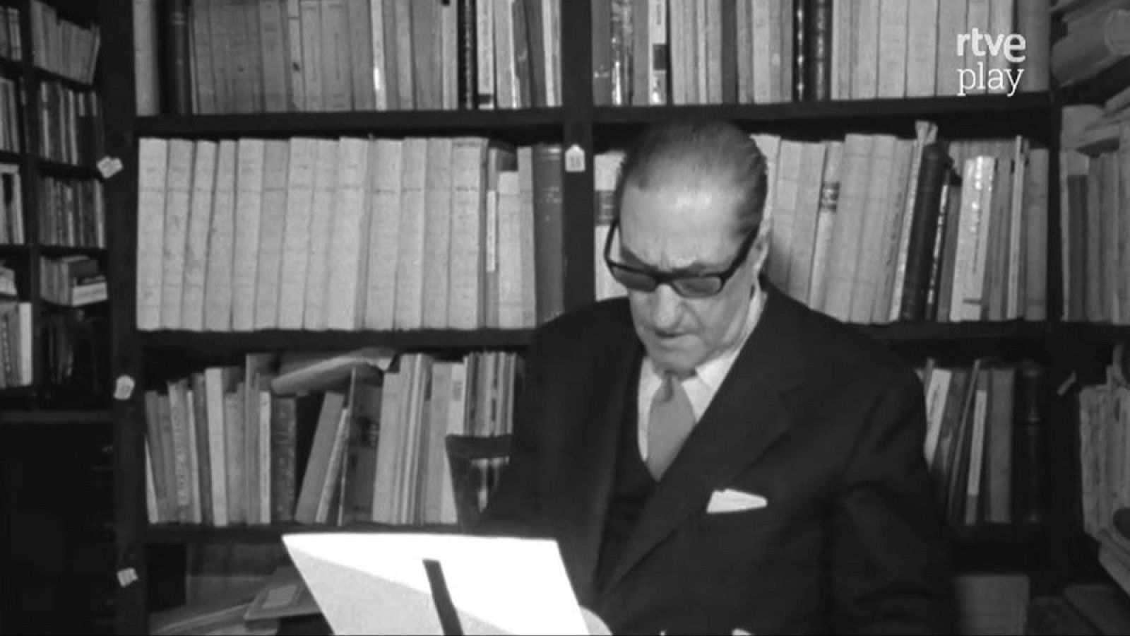 Un hombre con gafas, vestido con traje y corbata, lee un documento frente a una estantería llena de libros. El logo de "rtve play" se encuentra en la esquina superior derecha.