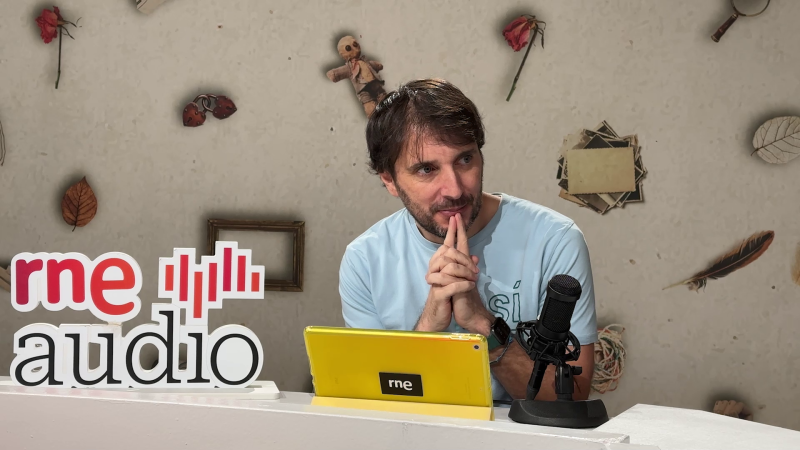 Un hombre con camiseta azul clara y barba incipiente está sentado frente a una mesa con un logo y una tablet. A su lado, un micrófono profesional. En la pared, objetos decorativos.