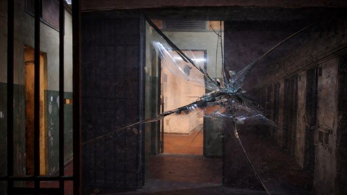 Un pasillo oscuro y deteriorado, con barrotes y una puerta abierta al fondo, muestra un ambiente sombrío y de abandono. Un cristal roto en primer plano añade un elemento de tensión a la escena.