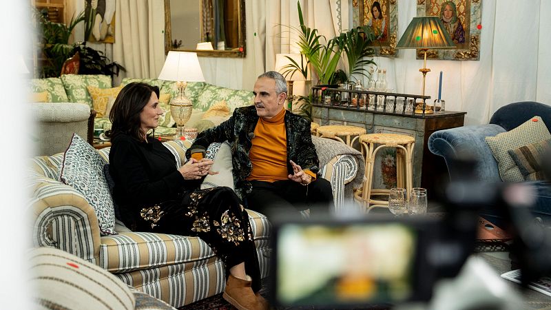 Dos personas, posiblemente en un estudio, posan sentadas en sofás. Una mujer viste de negro con detalles dorados y botines, y un hombre lleva un jersey naranja y una chaqueta estampada. El entorno está decorado con elementos como lámparas y cuadros.