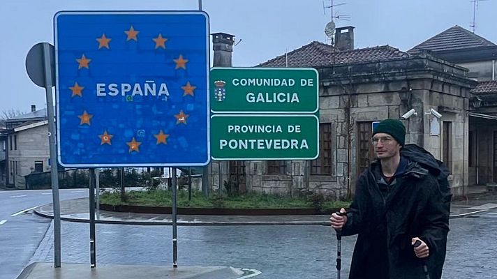 Un hombre con gafas y gorro, con bastones de senderismo, se encuentra cerca de señales de tráfico que indican la frontera española, la \"COMUNIDAD DE GALICIA\" y la \"PROVINCIA DE PONTEVEDRA\", en un día nublado.