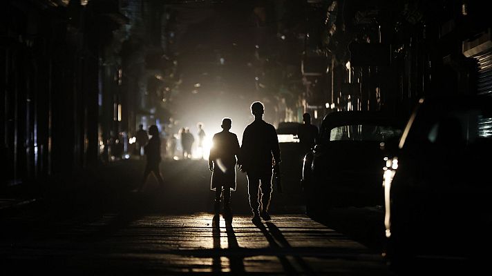 En una calle oscura, dos siluetas caminan hacia una luz brillante, proyectando sombras largas. Un coche estacionado y otras figuras difusas contribuyen a la atmósfera misteriosa de la escena.