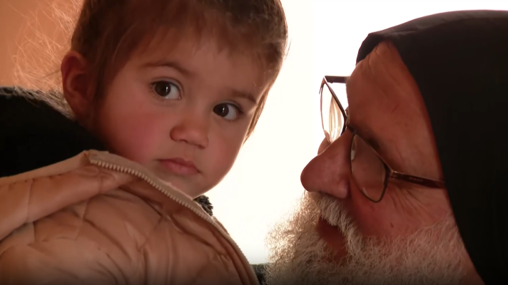 Un sacerdote maronita con gafas y barba blanca abraza a una niña pequeña que viste una chaqueta clara, ambos mirando a la cámara. La imagen podría representar la acogida de refugiados en un contexto religioso.
