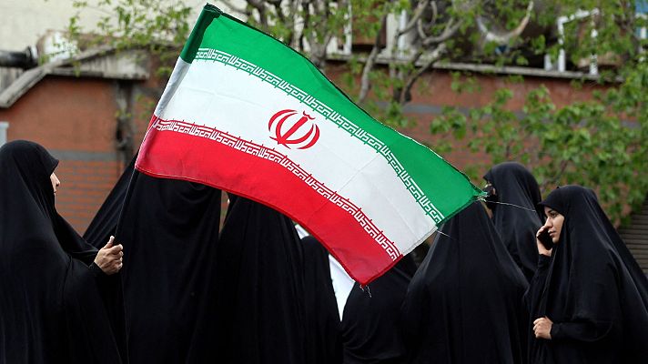 Mujeres iraníes con chador negro sostienen la bandera de Irán en un entorno urbano, posiblemente durante una manifestación. La bandera muestra las franjas verde, blanca y roja, con el emblema nacional en el centro.