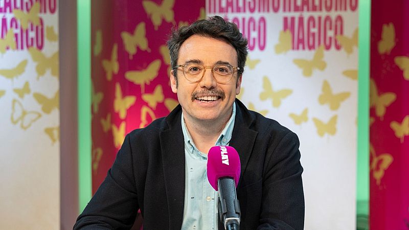 Un hombre con gafas y bigote, sonríe a la cámara, vestido con camisa clara y chaqueta oscura, frente a un fondo con diseño de mariposas y el texto "INFLU-REALISMO MÁGICO", con un micrófono rosa con el logo "PLAY".