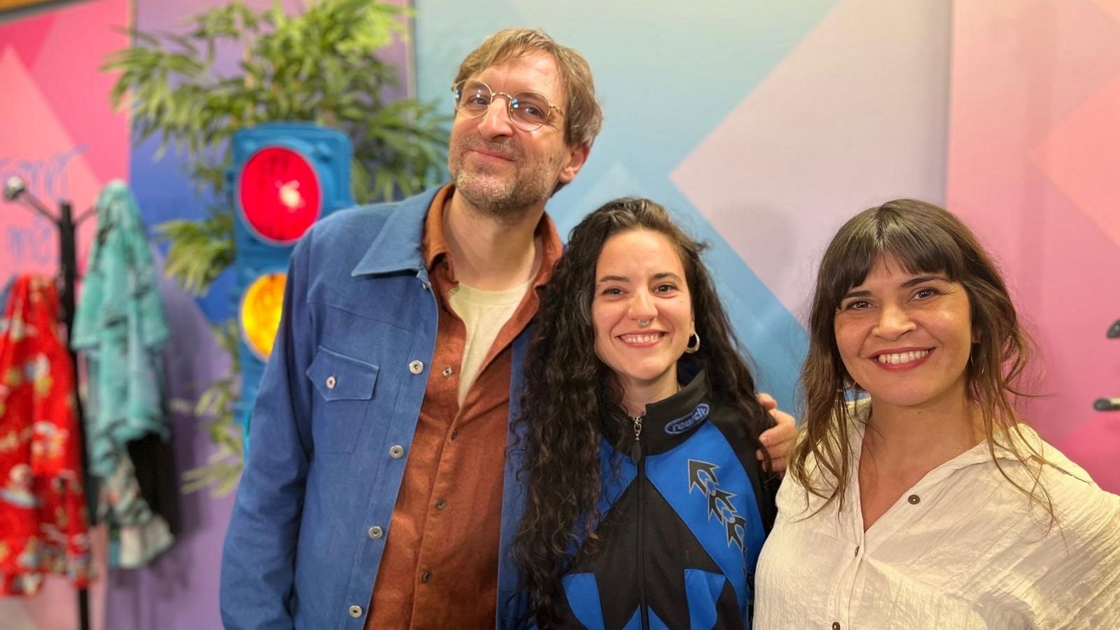 Tres personas posan frente a una pared decorada con diseños geométricos. Un hombre con gafas y chaqueta azul está a la izquierda, una mujer con cabello rizado y chaqueta azul y negra en el centro, y una mujer con flequillo y camisa blanca a la derecha.