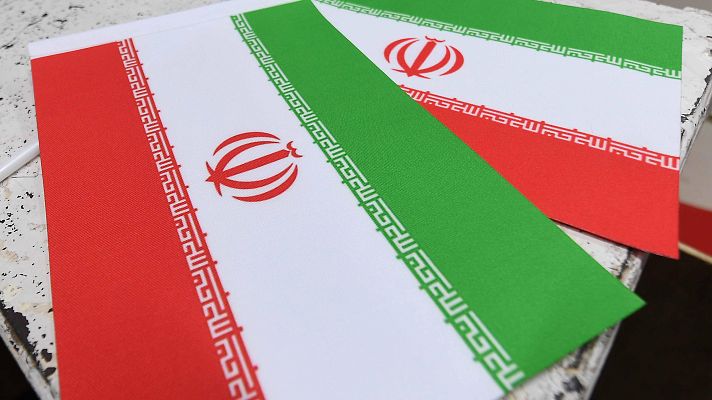 Dos banderas iraníes ondean, mostrando franjas horizontales rojas, blancas y verdes, con el emblema nacional rojo en el centro y escritura en los bordes.
