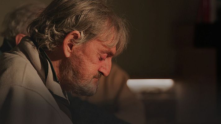 Un hombre de mediana edad, con cabello y barba canosos, aparece en primer plano con los ojos cerrados y una expresión serena, bajo una iluminación tenue. Viste una prenda clara.
