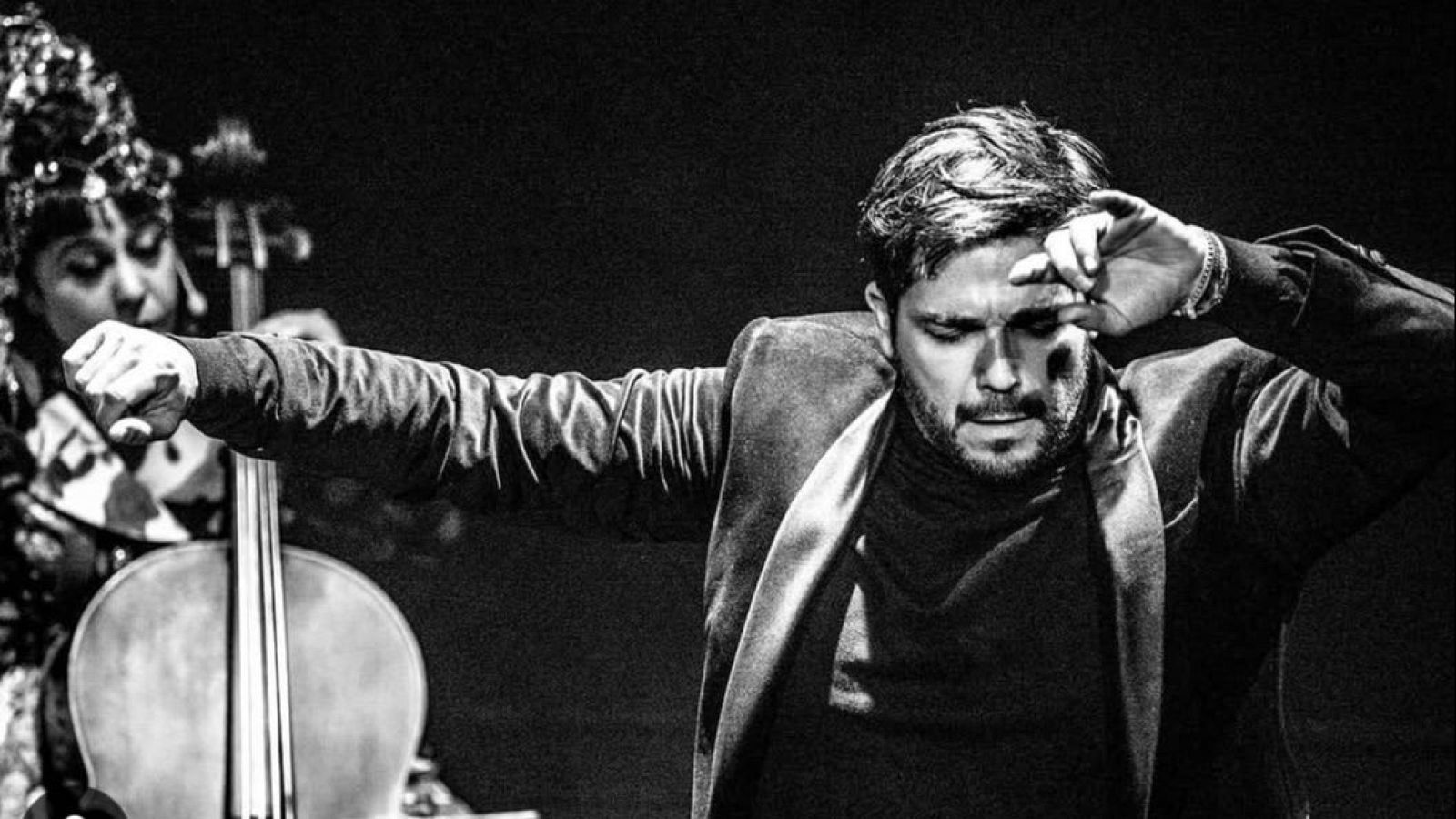 En una escena en blanco y negro, un hombre con traje oscuro y barba se lleva la mano a la frente, mientras una mujer con un tocado toca un instrumento musical de cuerda.