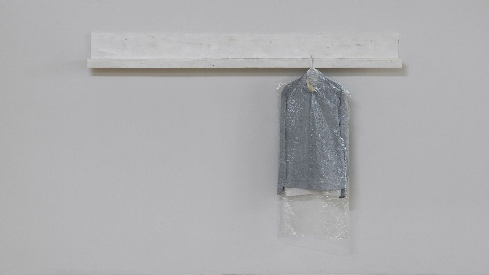 Una pared gris clara sirve de fondo a una estantería blanca, de la cual cuelga una percha metálica con una prenda de vestir gris protegida por una funda de plástico transparente.
