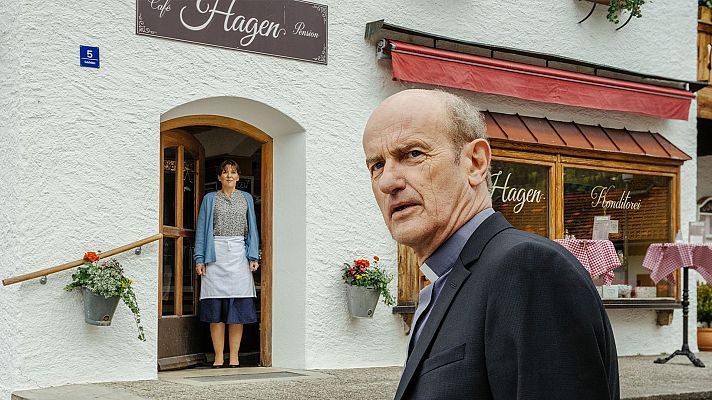 Michael Mendl como sacerdote frente a \"Cafe Hagen Pension\" y \"Hagen Konditorei\". Una mujer lo observa desde la entrada del establecimiento.
