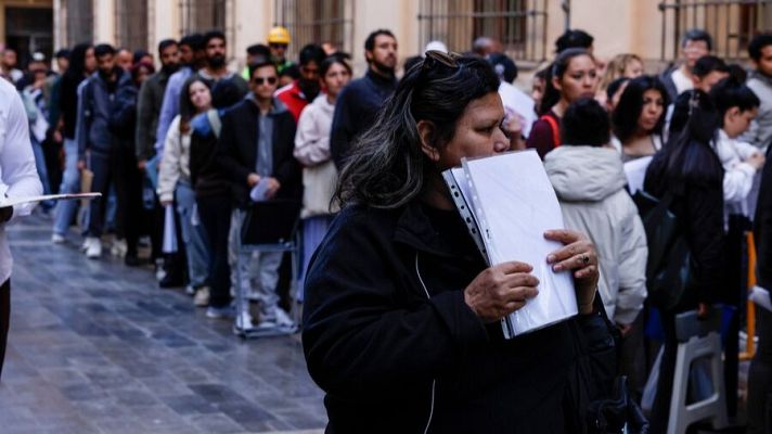 Persona con gafas y carpeta transparente en fila urbana. Edificio antiguo al fondo. Multitud diversa esperando.