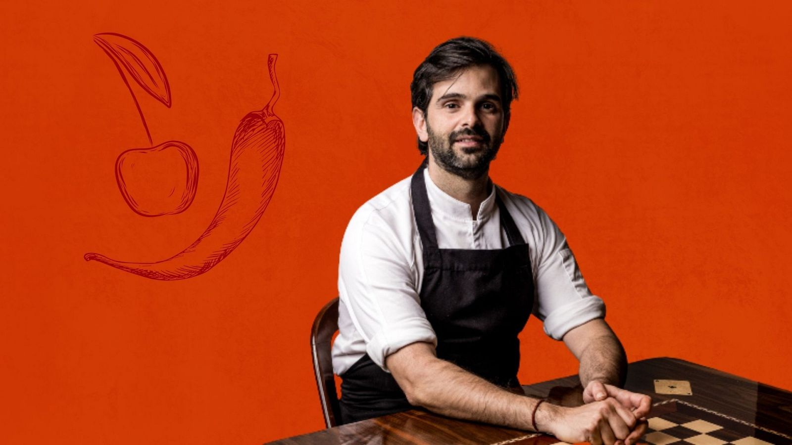 Chef Javi Estévez, camisa blanca, delantal negro, manos entrelazadas sobre mesa de madera oscura, fondo naranja rojizo con dibujos de cereza y guindilla.