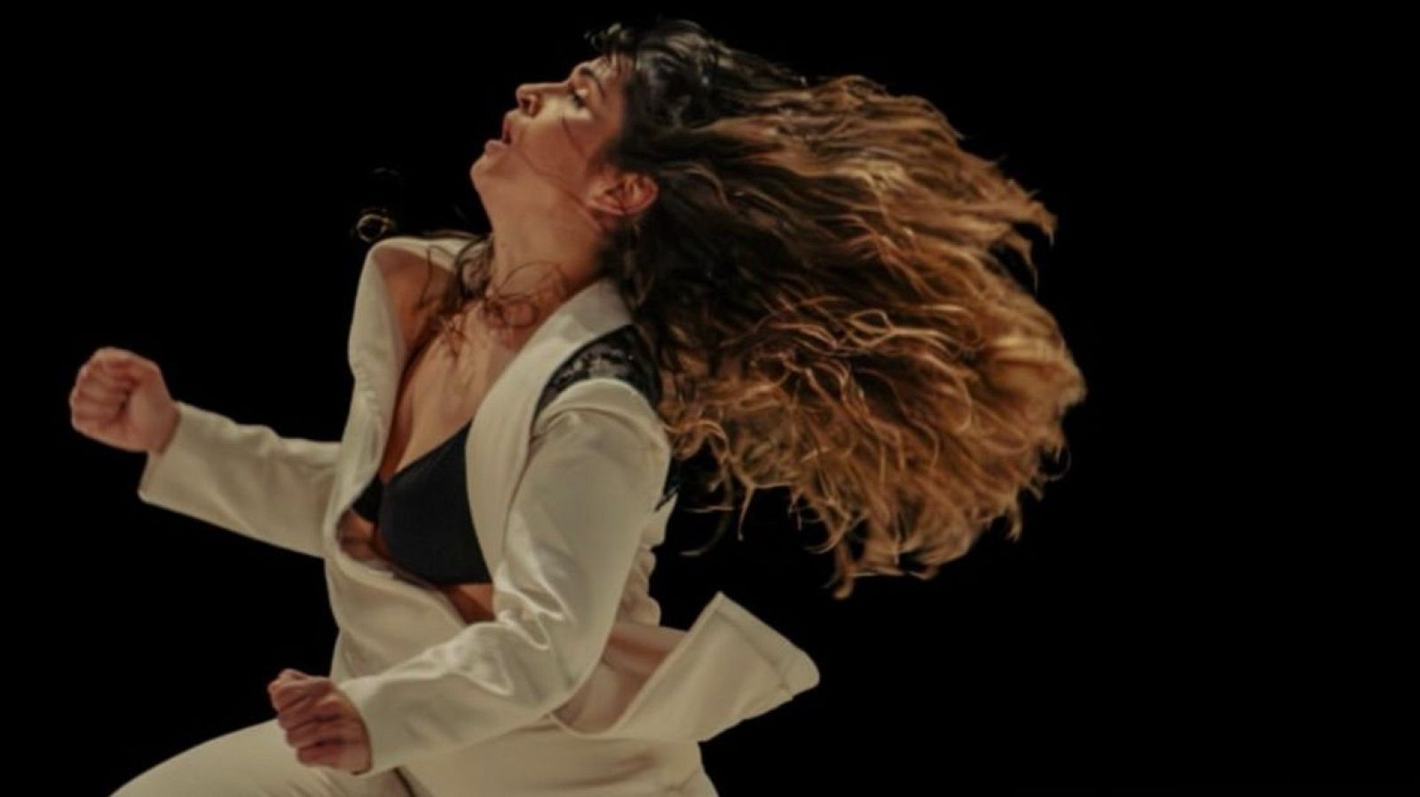 Mujer con cabello castaño rizado en movimiento, cabeza echada hacia atrás, boca abierta, chaqueta clara sobre top oscuro con detalles de encaje negro. Fondo negro.