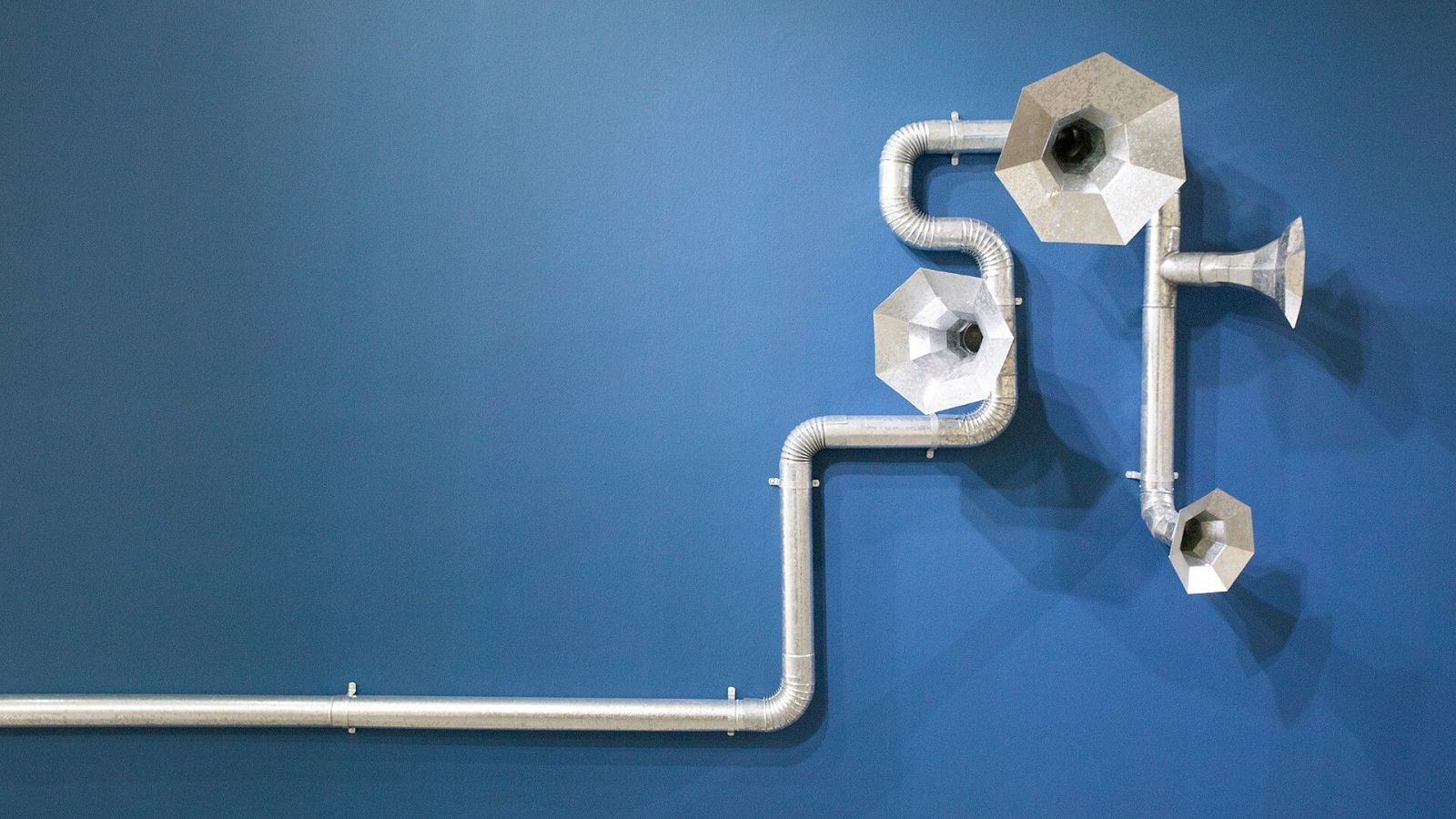 Instalación artística con tubos metálicos, conductos corrugados y cuatro bocinas (tres octogonales, una cónica) sobre pared azul cobalto. Estética industrial y moderna.