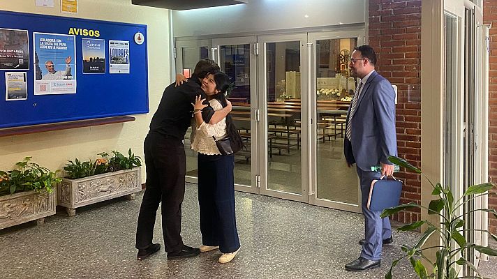 Tres personas en un interior, dos abrazándose, frente a un tablón de anuncios con \"VOLUNTARIADO\", \"VISITA DEL PAPA LEÓN XIV A MADRID\" (con imagen del Papa Francisco), \"Summer\" y \"Lourdes\".