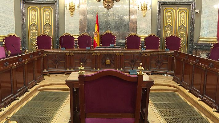 Sala institucional vacía en España, con mesa en \"U\", sillas rojas, bandera de España y paredes de mármol. Ambiente solemne.