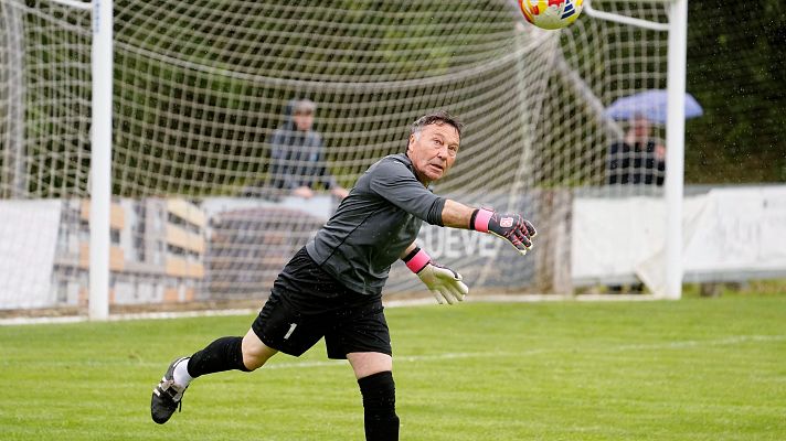 Portero con guantes rosas se lanza por un balón Adidas en un campo de fútbol. Al fondo, portería blanca, espectadores y un paraguas azul.