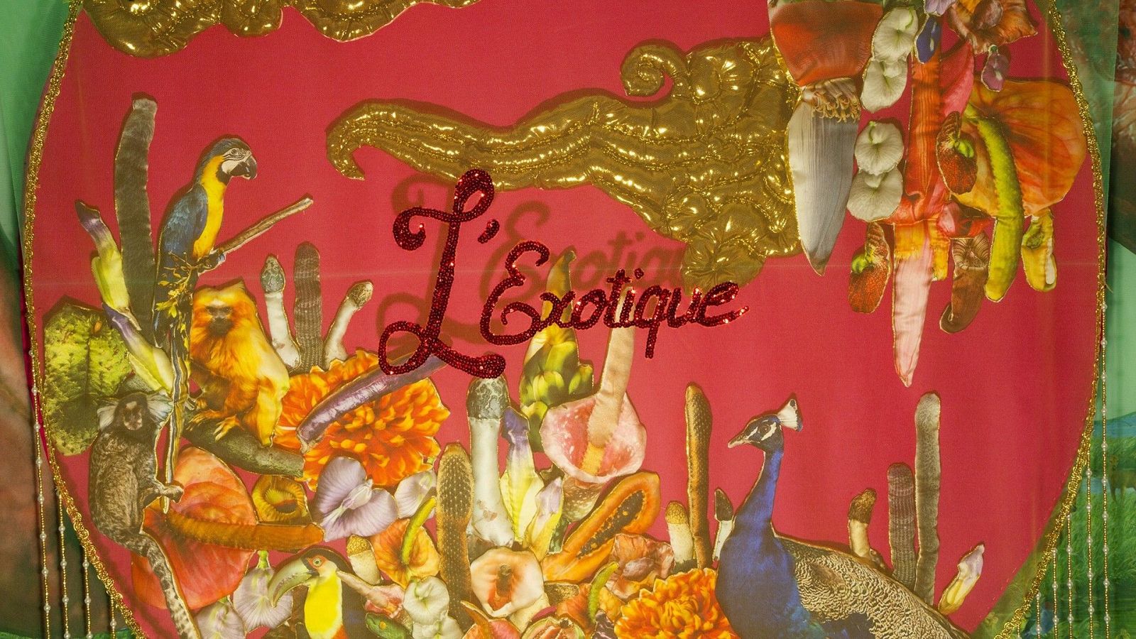 Composición tropical con fondo rojo, adornos dorados y el texto "L'Exotique". Presenta fauna exótica (guacamayos, titíes, pavo real) y flora exuberante (flores, cactus, frutas).