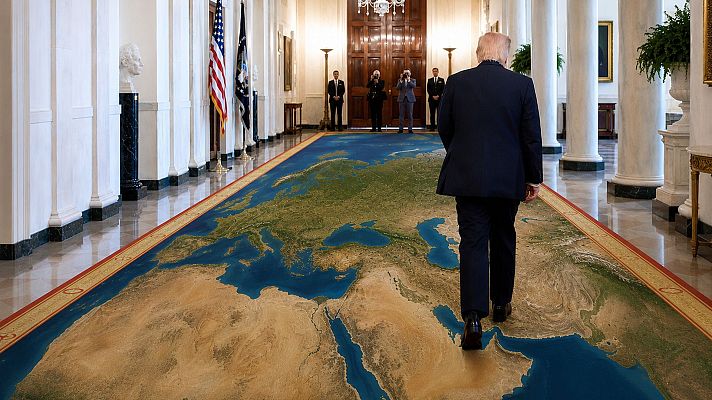Donald Trump camina por un pasillo con una alfombra de mapa mundial, flanqueado por columnas y decorado con banderas y un busto.