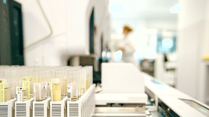Primer plano de tubos de ensayo etiquetados con códigos de barras y \"10 ml\", algunos con líquido amarillento, en un sistema automatizado de laboratorio. Al fondo, una persona con bata blanca.