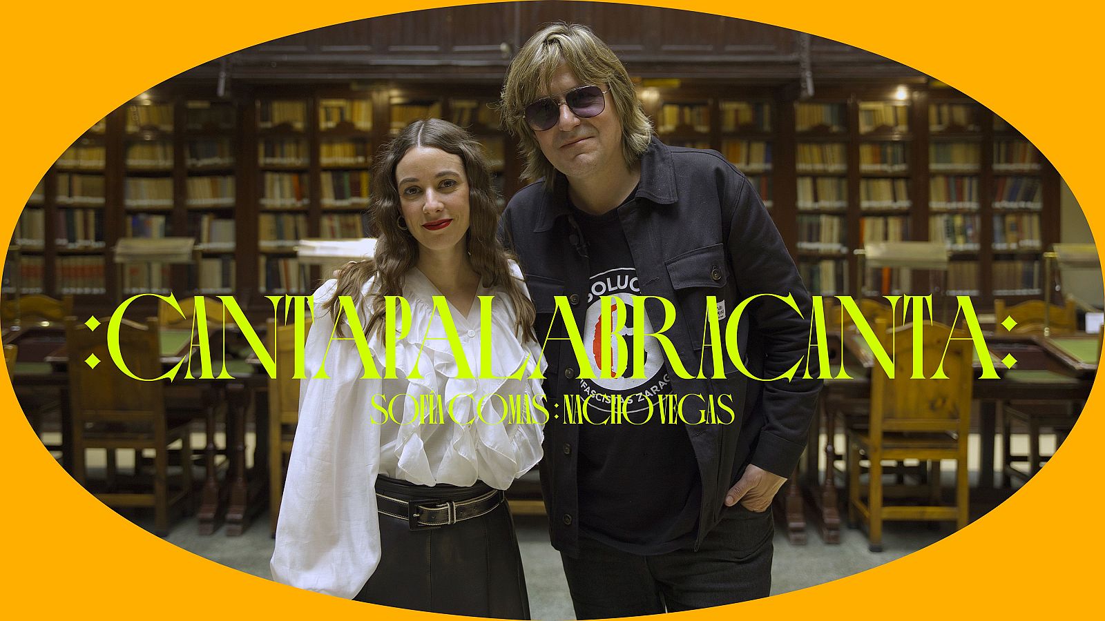 Nacho Vegas y una mujer posan en una biblioteca con el título ":CANTAPALABRACANTA:" y "SCRIPTOIUMS: NACHOVEGAS" superpuestos. Él lleva gafas de sol y una camiseta con logo; ella, blusa blanca y falda oscura.