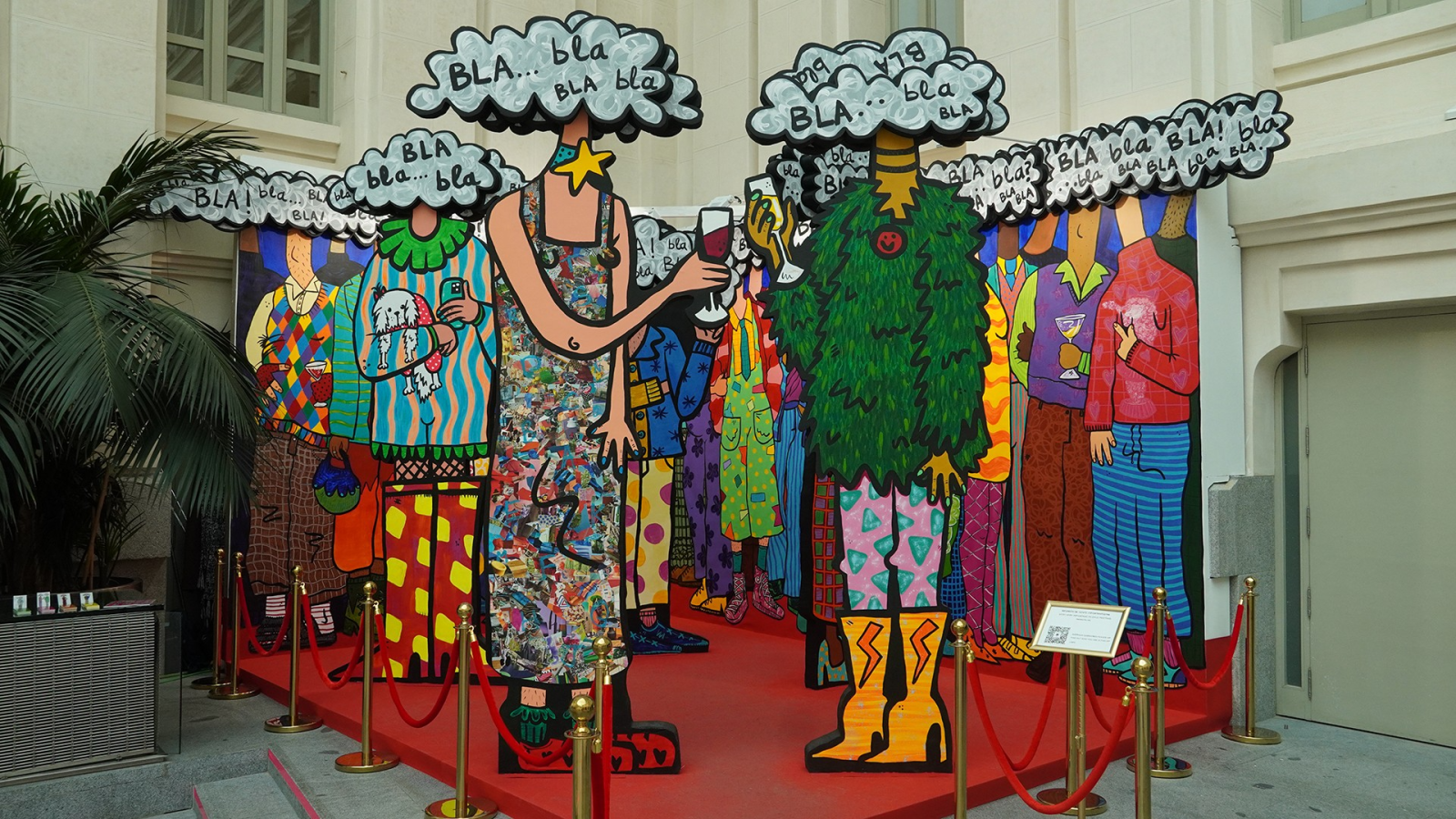 Instalación pop art con figuras estilizadas y cabezas de nube de diálogo "BLA... bla bla". Dos figuras centrales, una con vestido multicolor y otra con follaje, sostienen copas. Plataforma roja, postes dorados, palmera y edificio al fondo.