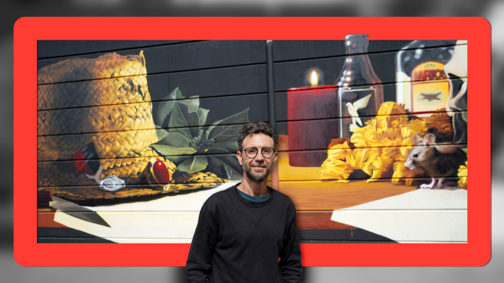 Hombre con barba y gafas frente a un mural de naturaleza muerta con sombrero, bayas, vela, botellas y flores con un ratón.