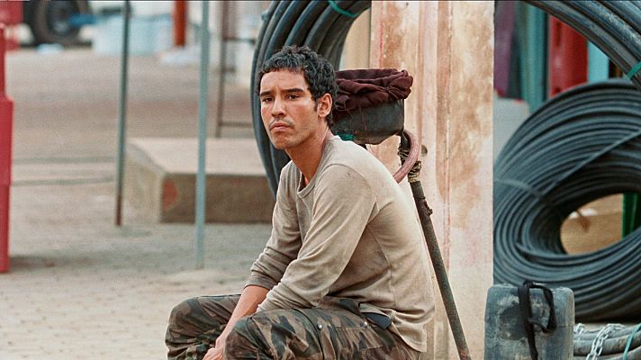Primer plano de Adam Bessa, actor de \"Harka\", con expresión seria. Viste camiseta beige y pantalones de camuflaje, en un entorno industrial con rollos de cable y pared texturizada.