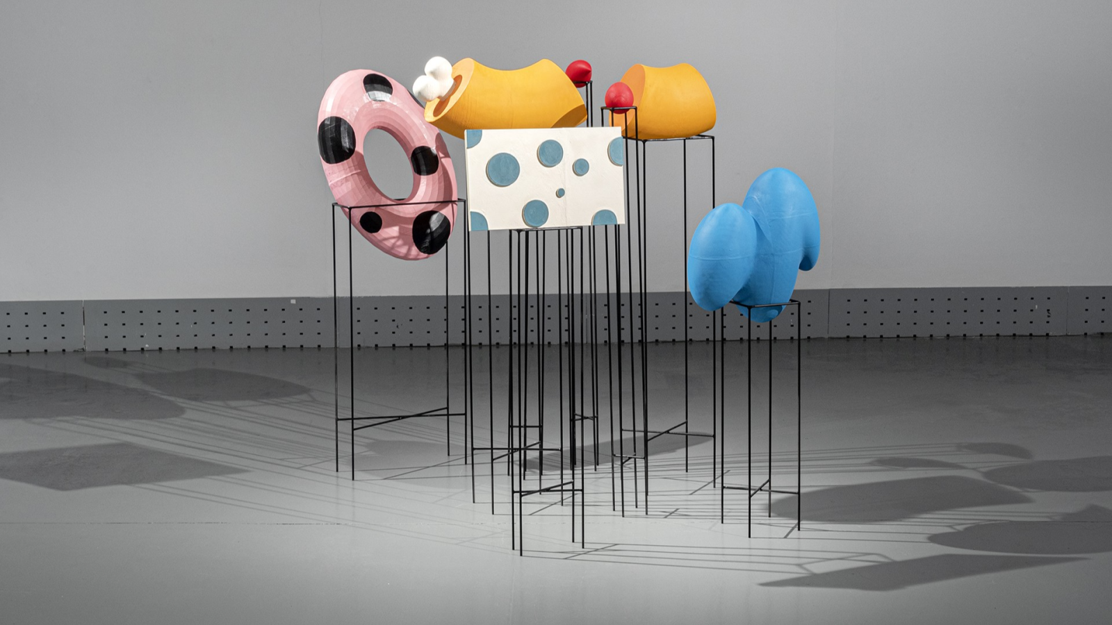 Instalación artística con objetos abstractos de colores (rosa, amarillo, rojo, azul) y formas variadas, suspendidos por varillas metálicas negras sobre un suelo gris.
