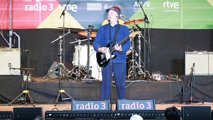 Carlangas, músico español, actúa en directo con guitarra y micrófono, luciendo un conjunto azul y gorra. El escenario está decorado con logos de Radio 3, RTVE y apoyo institucional.