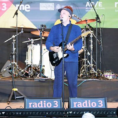 Carlangas, músico español, actúa en directo con guitarra y micrófono, luciendo un conjunto azul y gorra. El escenario está decorado con logos de Radio 3, RTVE y apoyo institucional.