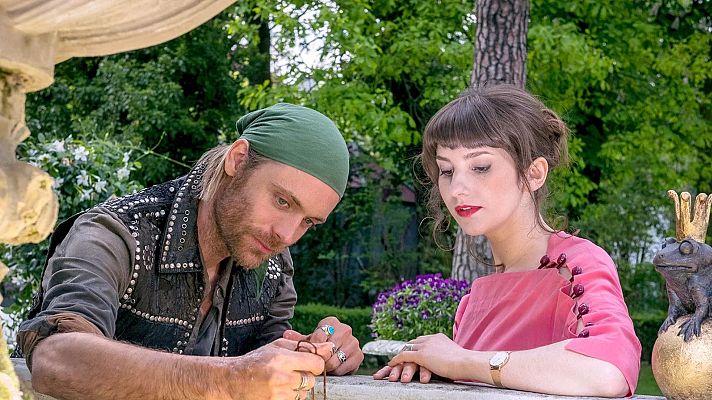 Hombre con bandana verde y mujer con labios rojos observan un objeto en un jardín. Detalle en manos y vestimenta. Fondo de vegetación y flores moradas.