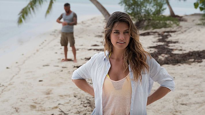 Blanca Suárez en una playa tropical, con camisa azul abierta sobre bikini amarillo, manos en caderas, sonriendo a cámara. Al fondo, un hombre en la orilla.