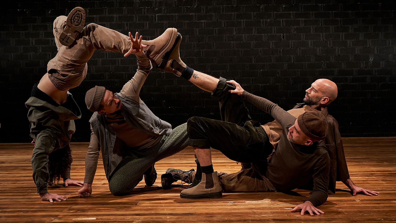 Cuatro personas en una pose acrobática sobre un escenario de madera con fondo de ladrillos. Se observa una compleja interacción de cuerpos en equilibrio y apoyo mutuo.