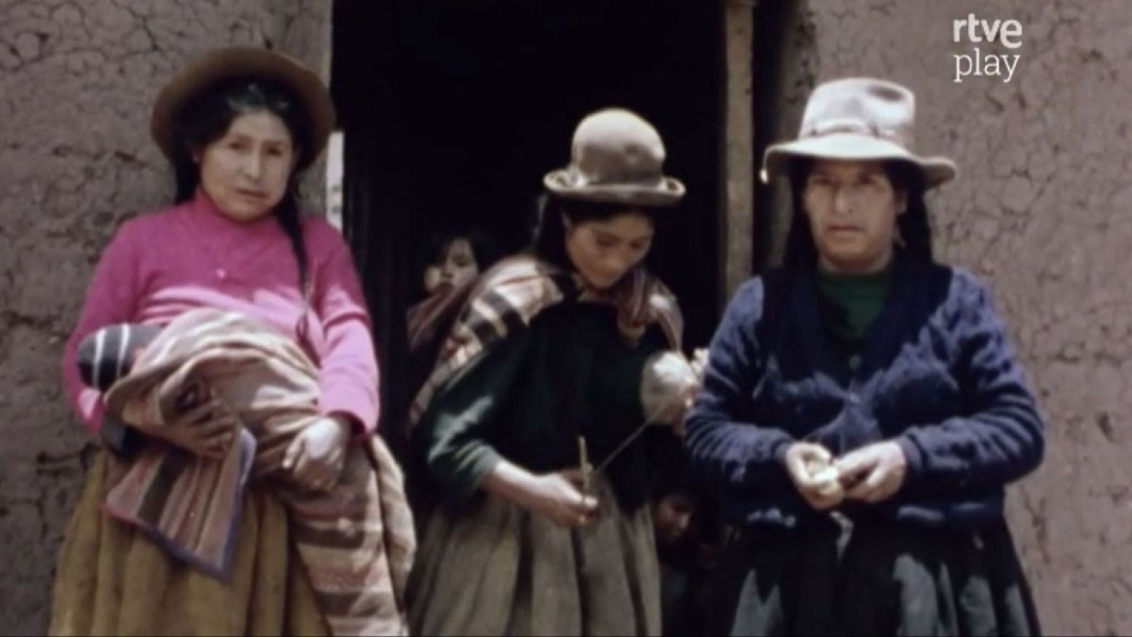 Mujeres Quechuas vestidas tradicionalmente, una hilando lana, posan frente a pared de adobe con niños detrás. Logo de rtve play visible.