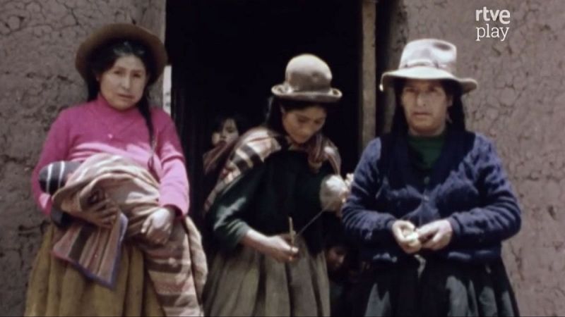Mujeres Quechuas vestidas tradicionalmente, una hilando lana, posan frente a pared de adobe con ni�os detr�s. Logo de rtve play visible.