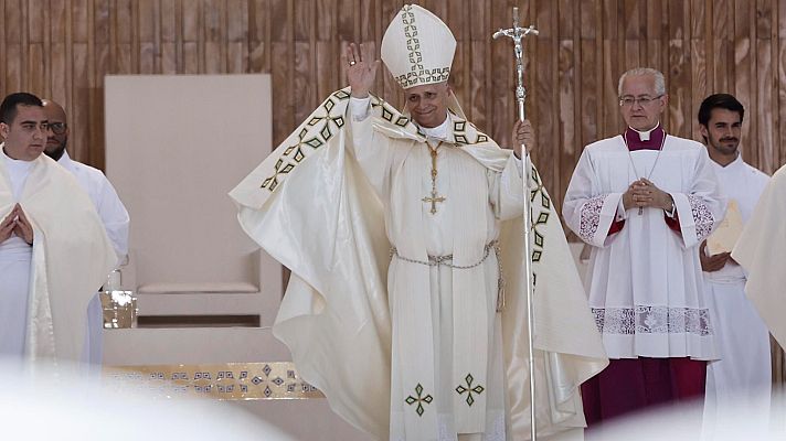 El Papa León XIV, con mitra y báculo, oficia una ceremonia. A su izquierda, un joven clérigo; a su derecha, un hombre mayor con gafas y sotana morada. Fondo de madera y panel blanco.