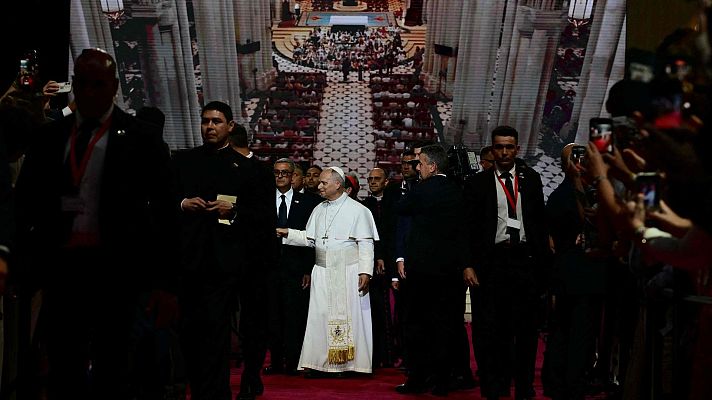 El Papa Francisco avanza por un pasillo alfombrado de rojo, acompañado por hombres de traje, mientras el público documenta el evento con sus teléfonos.