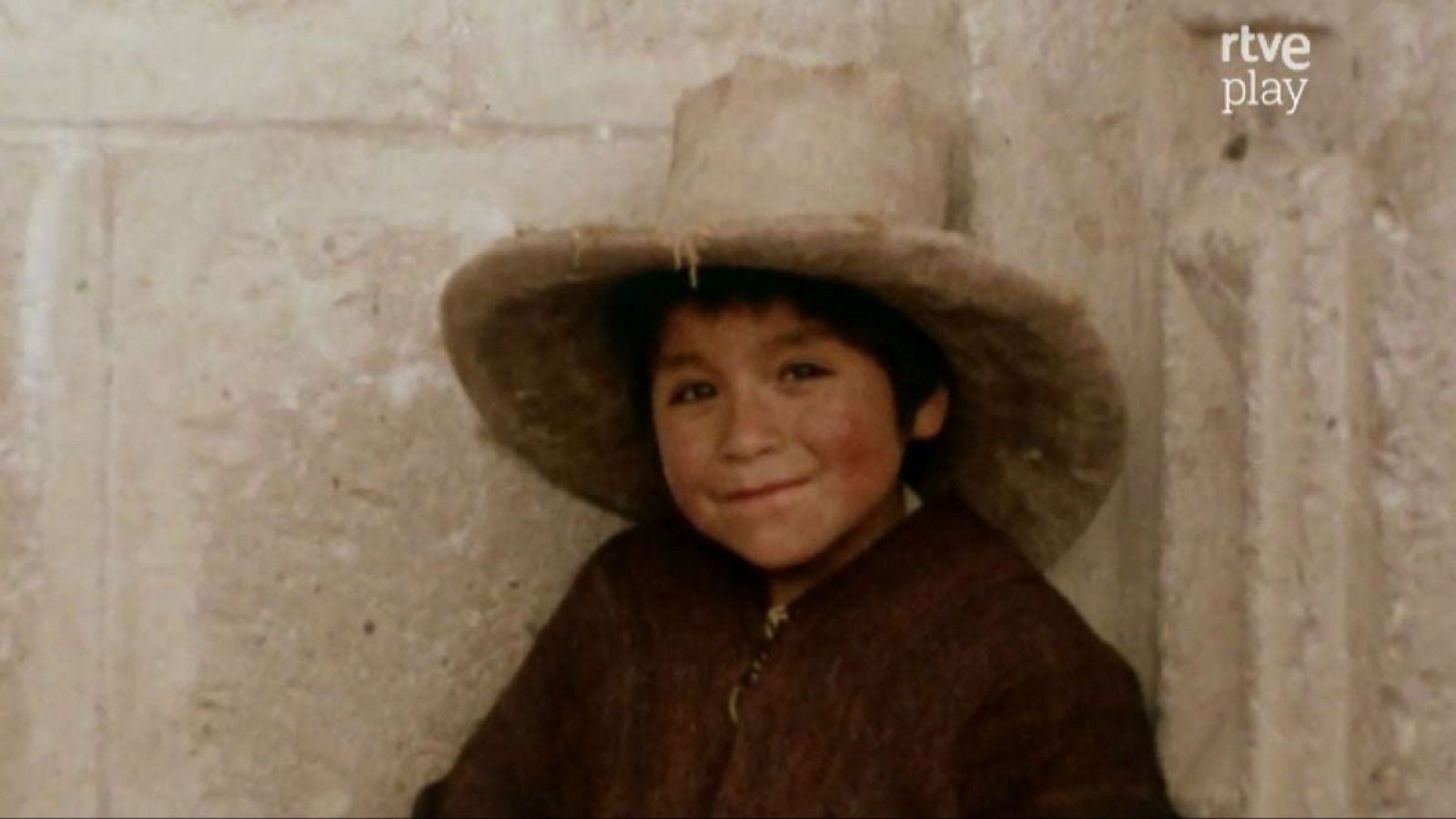 Niño de rasgos andinos, con sombrero y abrigo oscuro, sonríe inocentemente a cámara. Fondo de pared rústica. Logo "rtve play" visible.