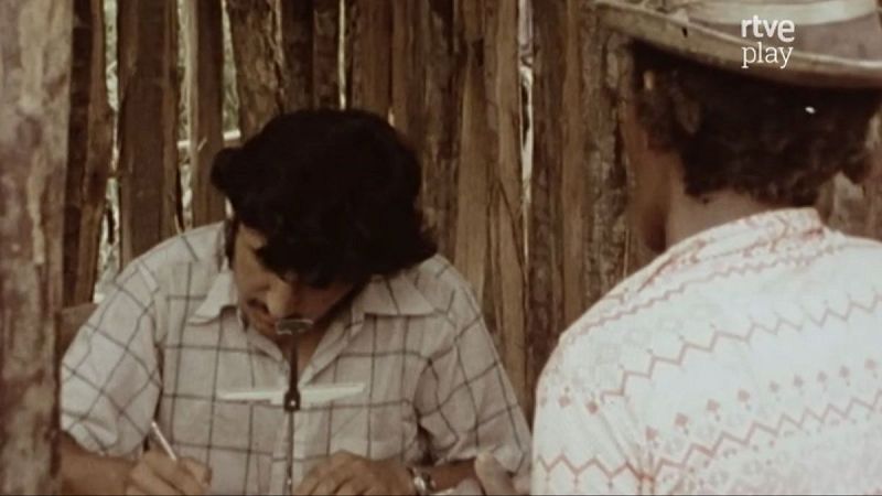 Hombres en entorno r�stico con paredes de madera. Uno escribe/dibuja con gorra y camisa a cuadros, el otro observa con sombrero. Logo "rtve play" visible.
